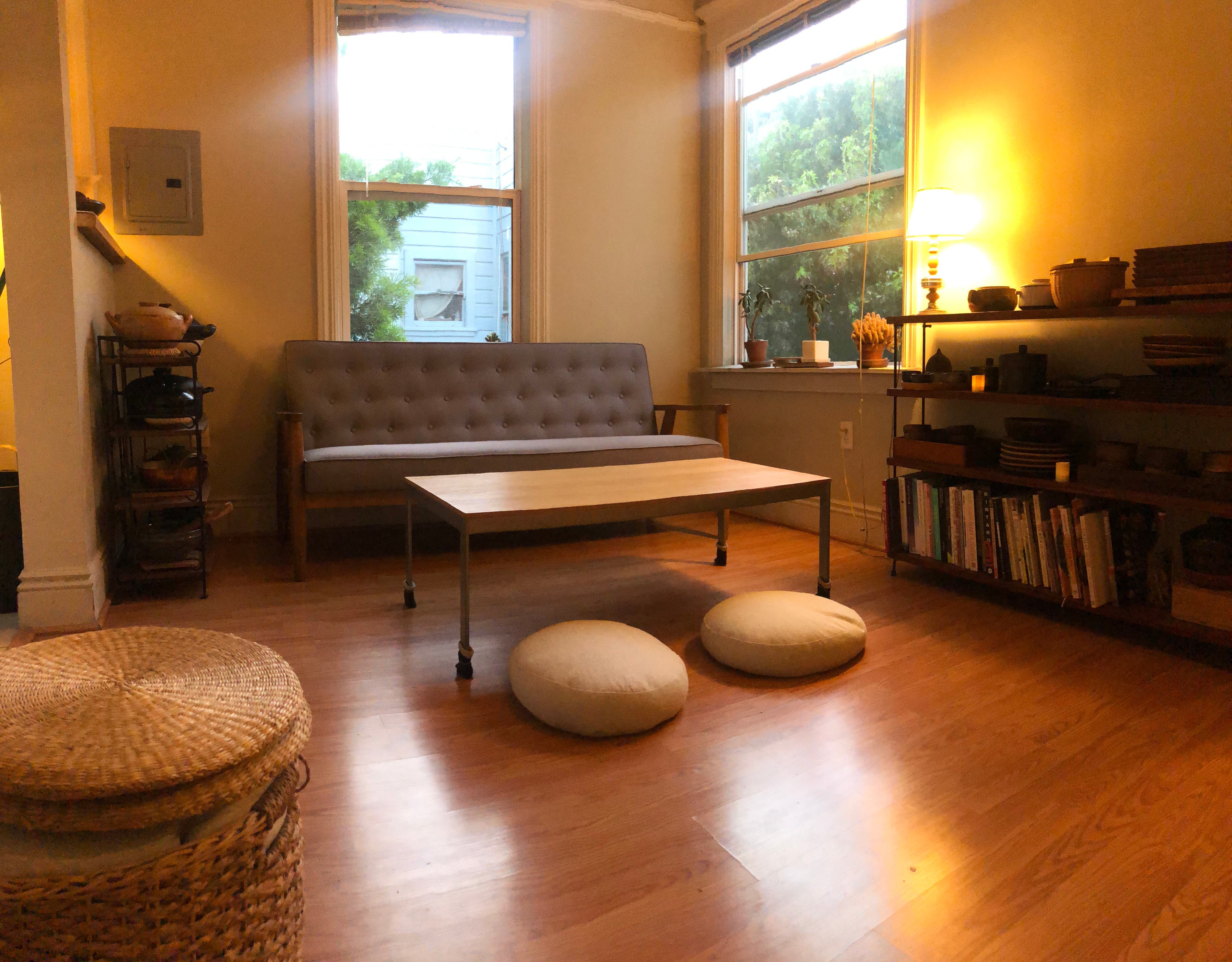 The room where dinner happens — a small living room in evening light, with a grey sofa, a low wooden table set between two round floor cushions, tall windows onto the trees, and shelves of books and pottery.